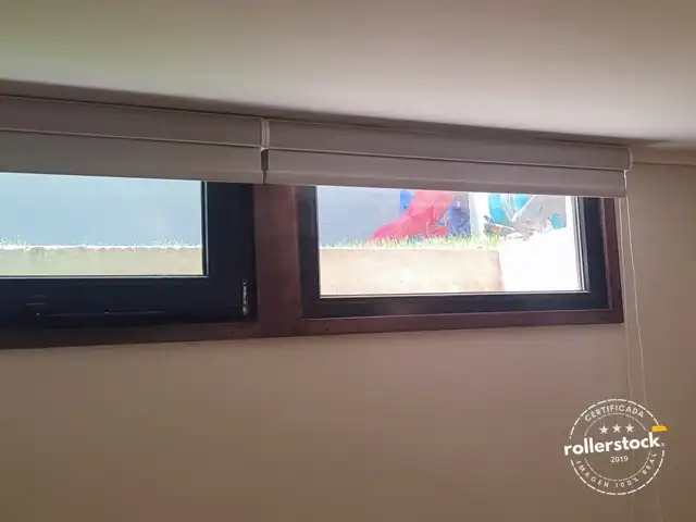 Dos cortinas roller blackout con cenefa color castaño, recogidas, instaladas en un ventanal pequeño.